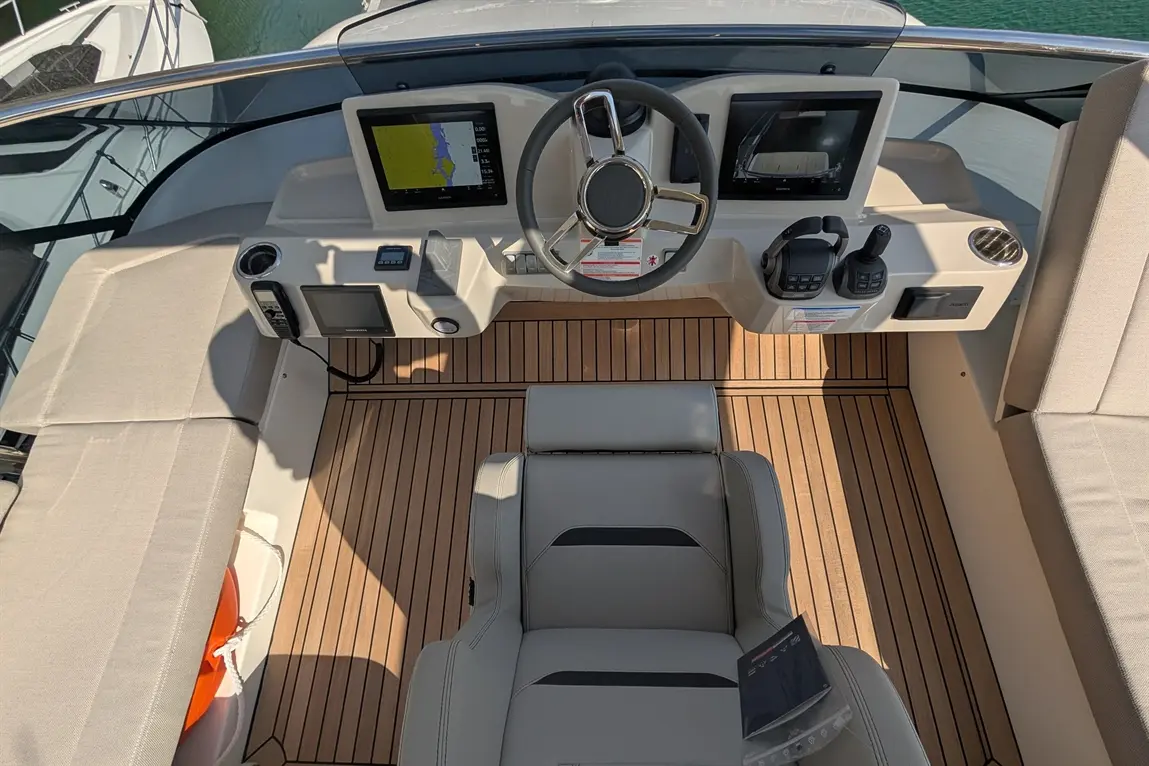 View from above a boat's helm featuring a steering wheel, navigation screens, and a grey captain's seat on wooden decking.